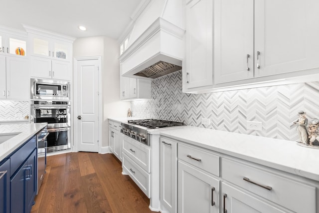 kitchen with stainless steel appliances, white cabinets, blue cabinetry, custom exhaust hood, and glass insert cabinets