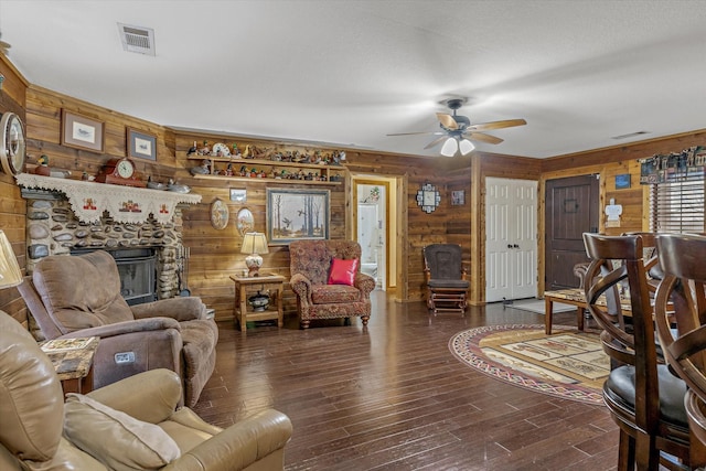 living area featuring a stone fireplace, wood walls, wood finished floors, visible vents, and a ceiling fan