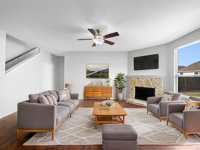 living area with ceiling fan, a stone fireplace, a textured ceiling, and wood finished floors