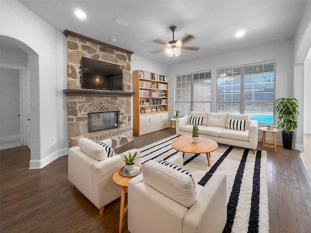 living area with arched walkways, a stone fireplace, wood finished floors, a ceiling fan, and baseboards