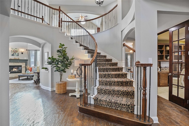 stairway with wood finished floors, a glass covered fireplace, ceiling fan, arched walkways, and a towering ceiling