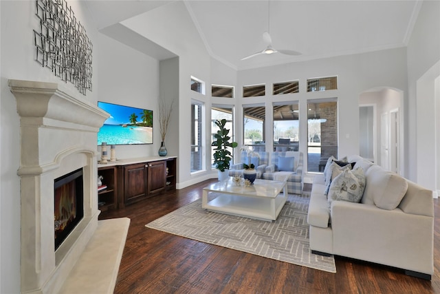 living area with a warm lit fireplace, high vaulted ceiling, ceiling fan, ornamental molding, and dark wood-style floors