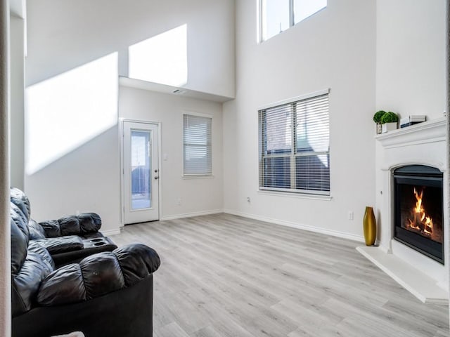 living room with a lit fireplace, a towering ceiling, light wood-style flooring, and baseboards