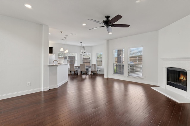 unfurnished living room with dark wood finished floors, recessed lighting, a warm lit fireplace, baseboards, and ceiling fan with notable chandelier