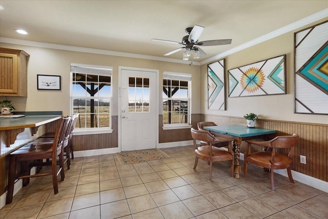 dining space with light tile patterned floors, wooden walls, wainscoting, ceiling fan, and ornamental molding