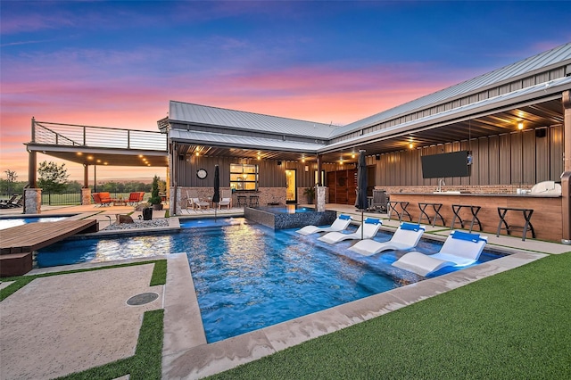 pool at dusk with a patio area, an outdoor pool, outdoor dry bar, and fence