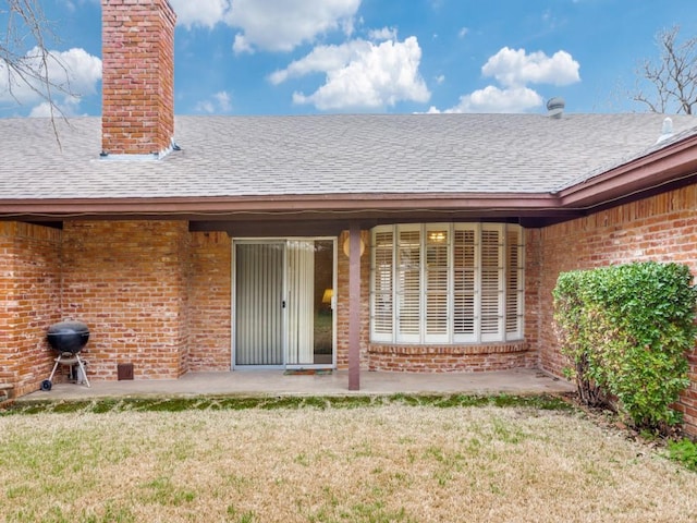 view of exterior entry with brick siding, roof with shingles, a chimney, and a lawn