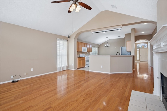 unfurnished living room with arched walkways, a tiled fireplace, lofted ceiling, light wood-type flooring, and ceiling fan with notable chandelier