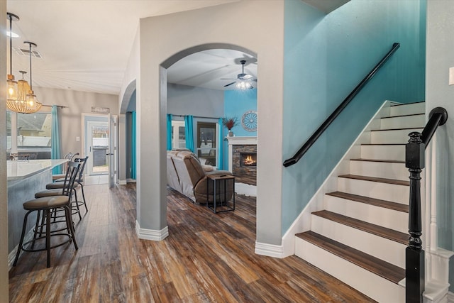 interior space with arched walkways, a fireplace, stairway, dark wood-type flooring, and ceiling fan