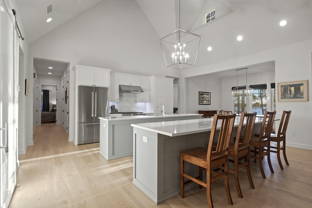 kitchen featuring under cabinet range hood, high end fridge, light wood finished floors, a large island with sink, and an inviting chandelier