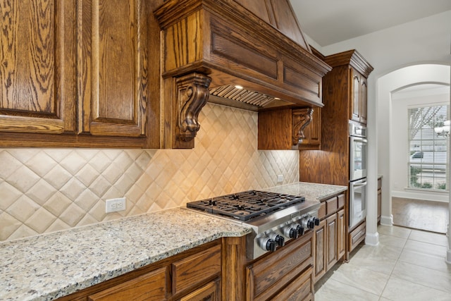 kitchen with arched walkways, light stone counters, stainless steel appliances, custom exhaust hood, and tasteful backsplash