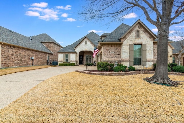 french country home featuring central air condition unit, a shingled roof, brick siding, stone siding, and a front yard