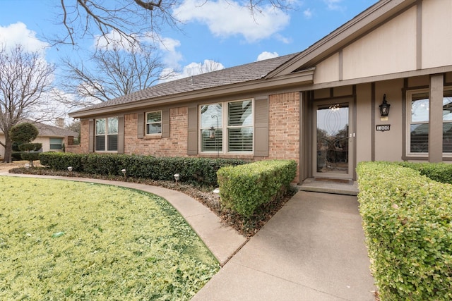 doorway to property featuring a shingled roof, a lawn, and brick siding