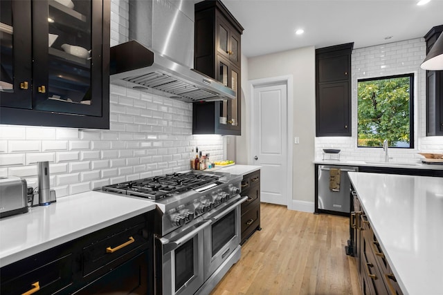 kitchen featuring light wood-style flooring, a sink, light countertops, wall chimney range hood, and appliances with stainless steel finishes