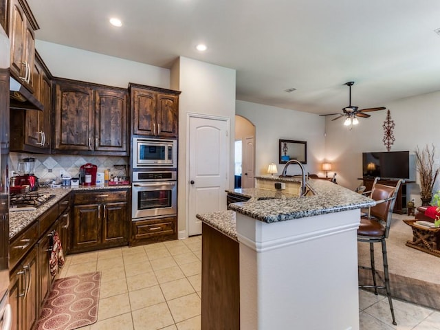 kitchen featuring arched walkways, stainless steel appliances, stone counters, a kitchen bar, and backsplash