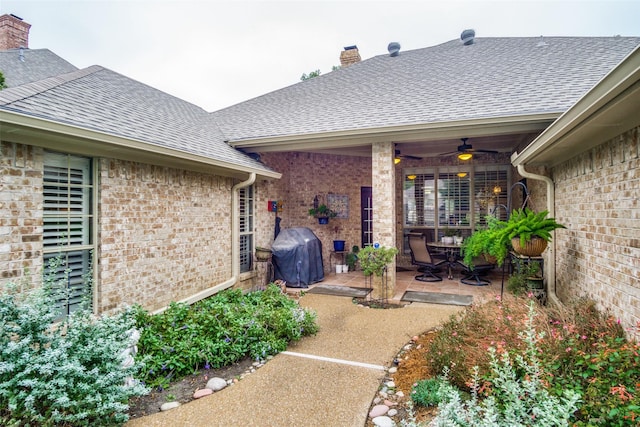 property entrance featuring a patio, brick siding, a ceiling fan, roof with shingles, and a chimney