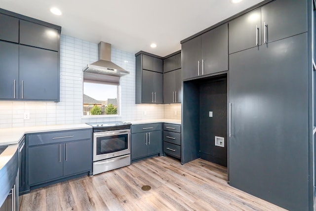 kitchen featuring stainless steel electric stove, light wood-style floors, light countertops, wall chimney range hood, and decorative backsplash