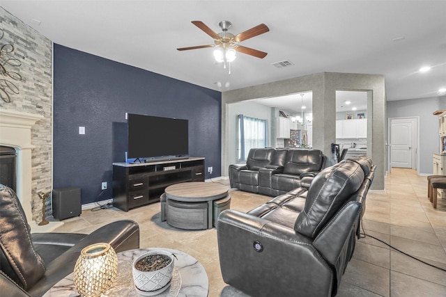 living room featuring light tile patterned floors, visible vents, baseboards, a stone fireplace, and ceiling fan with notable chandelier