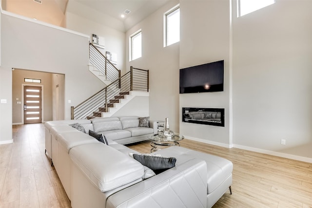 living area featuring stairway, a towering ceiling, a glass covered fireplace, light wood-type flooring, and baseboards