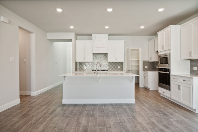 kitchen featuring black oven, stainless steel microwave, light wood-style flooring, and an island with sink