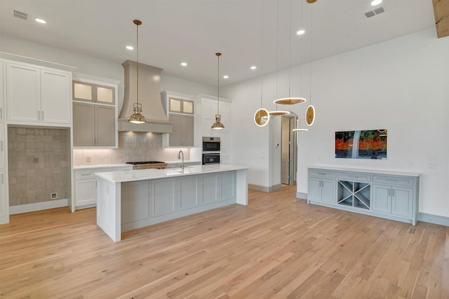 kitchen featuring visible vents, an island with sink, custom range hood, light countertops, and light wood-type flooring