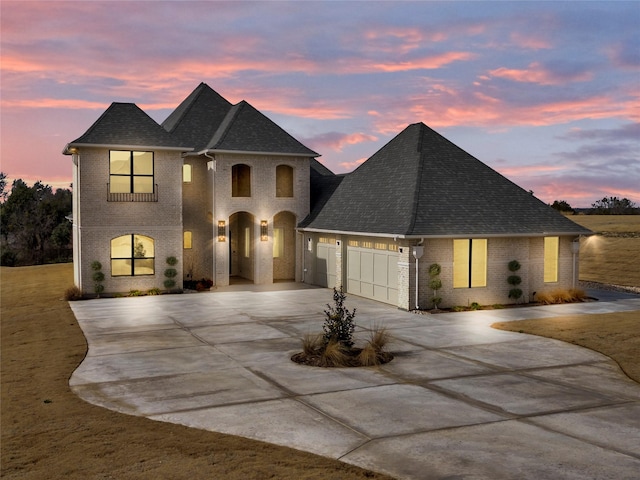 french country style house with a garage, driveway, brick siding, and roof with shingles