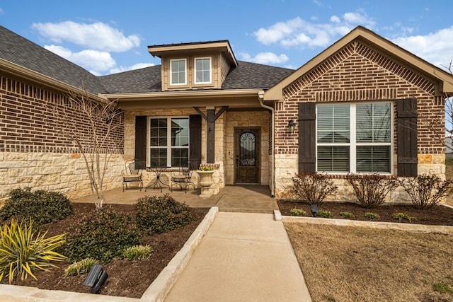 view of exterior entry with covered porch, stone siding, brick siding, and roof with shingles