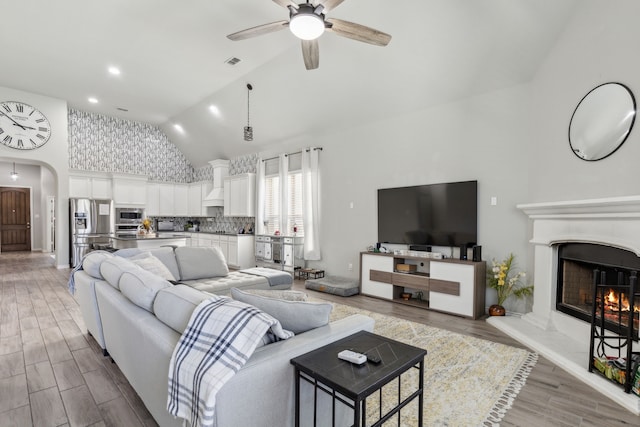 living room featuring arched walkways, a ceiling fan, visible vents, light wood-style floors, and a lit fireplace