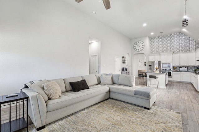 living room with light wood-type flooring, arched walkways, ceiling fan, and high vaulted ceiling