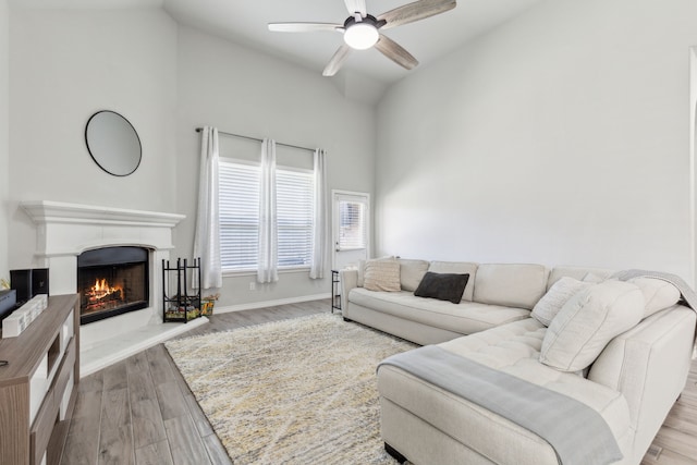 living area featuring ceiling fan, wood finished floors, high vaulted ceiling, a lit fireplace, and baseboards