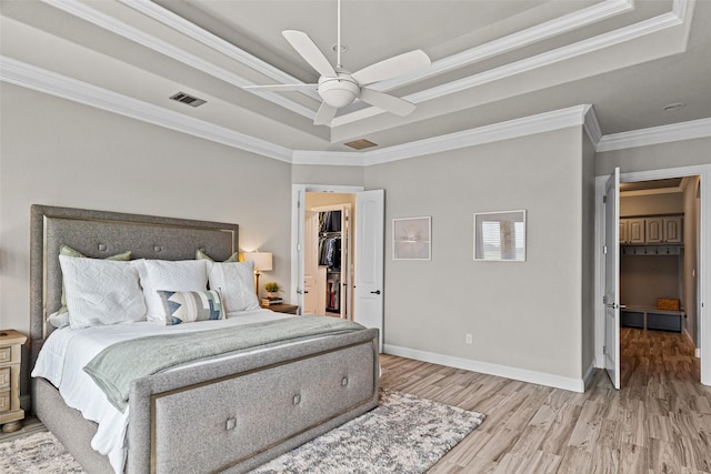 bedroom with a walk in closet, a raised ceiling, light wood-style flooring, ornamental molding, and baseboards