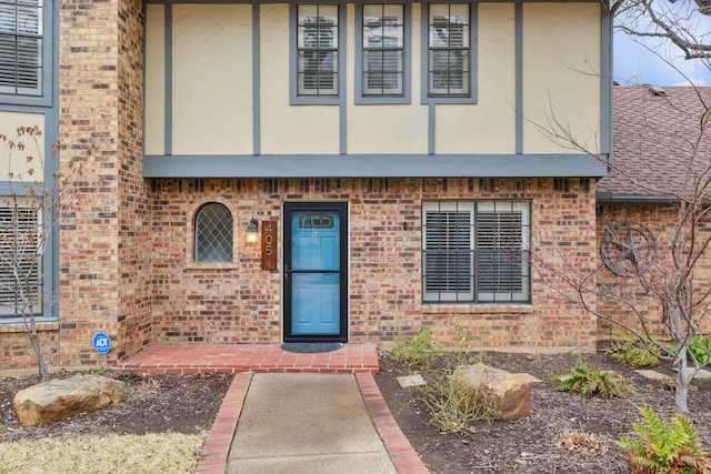 entrance to property featuring brick siding, roof with shingles, and stucco siding