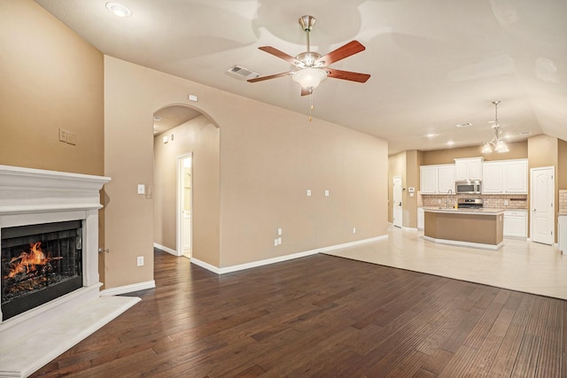 unfurnished living room with visible vents, a ceiling fan, a warm lit fireplace, wood finished floors, and arched walkways