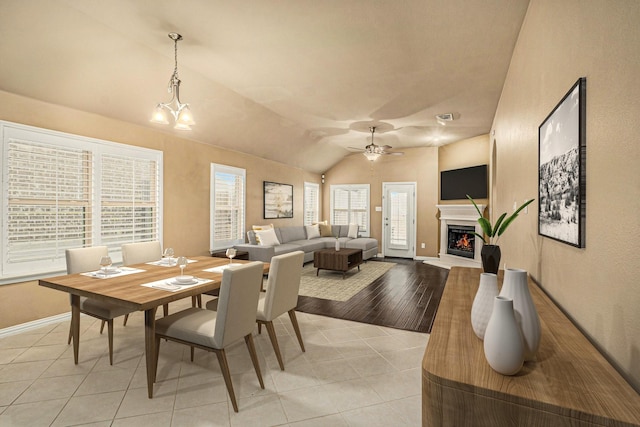 dining space featuring lofted ceiling, ceiling fan with notable chandelier, a glass covered fireplace, light tile patterned flooring, and baseboards