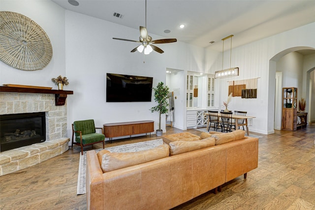 living room with arched walkways, visible vents, a ceiling fan, a stone fireplace, and wood finished floors