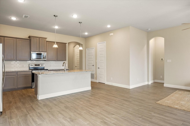 kitchen with arched walkways, a kitchen island with sink, appliances with stainless steel finishes, backsplash, and light wood finished floors