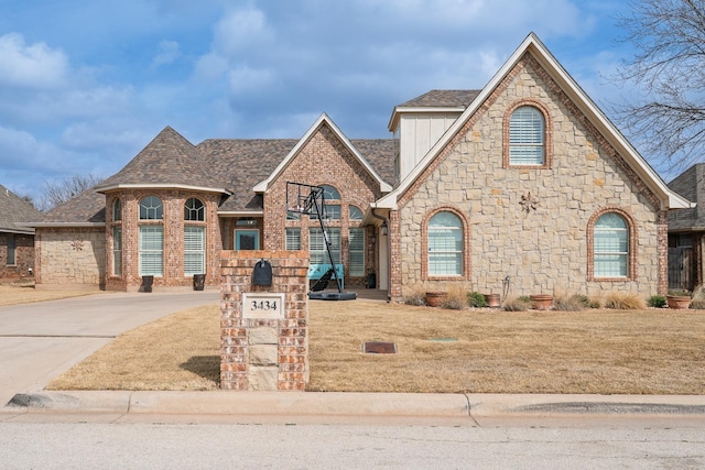 french country inspired facade with brick siding, roof with shingles, concrete driveway, and a front yard