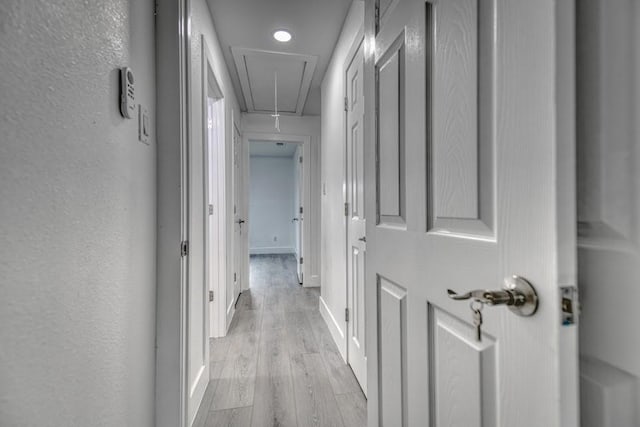 hall featuring a textured wall, light wood finished floors, attic access, and baseboards