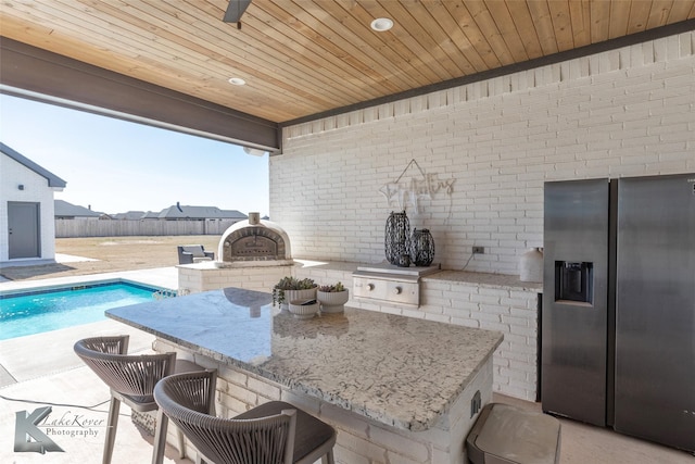 kitchen with light stone counters, wooden ceiling, stainless steel fridge, and brick wall