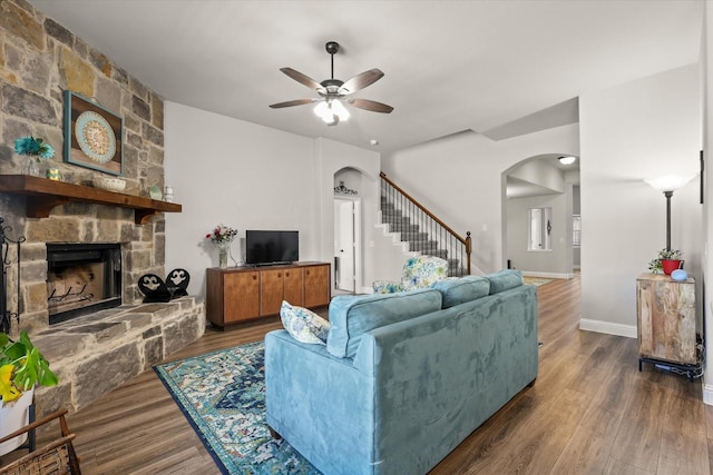 living area with arched walkways, ceiling fan, stairway, wood finished floors, and a stone fireplace