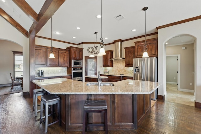 kitchen featuring arched walkways, appliances with stainless steel finishes, a large island with sink, wall chimney range hood, and a sink