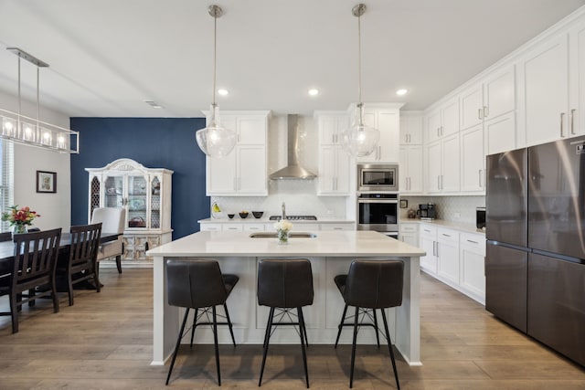 kitchen with a kitchen island with sink, wood finished floors, wall chimney range hood, appliances with stainless steel finishes, and decorative backsplash