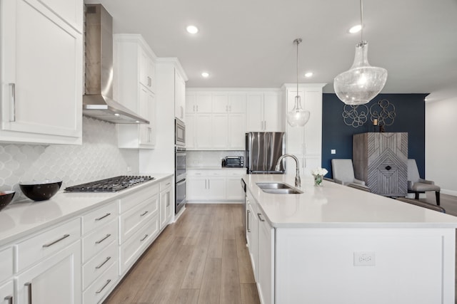 kitchen with white cabinets, light wood-style flooring, stainless steel appliances, wall chimney range hood, and a sink