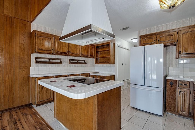 kitchen featuring tile countertops, black electric stovetop, custom exhaust hood, visible vents, and freestanding refrigerator