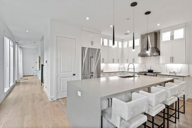 kitchen with a center island with sink, wall chimney exhaust hood, appliances with stainless steel finishes, light wood-type flooring, and a sink