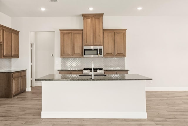 207 Cortese Ln, Alvarado, TX 76009 - kitchen featuring light wood-style floors, an island with sink, dark stone countertops, stainless steel microwave, and decorative backsplash
