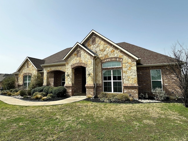 french country style house featuring stone siding, roof with shingles, a front yard, and brick siding