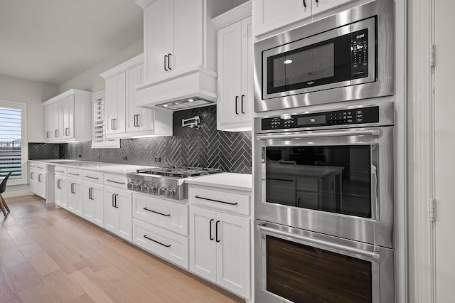 kitchen featuring light wood finished floors, light countertops, custom range hood, decorative backsplash, and appliances with stainless steel finishes