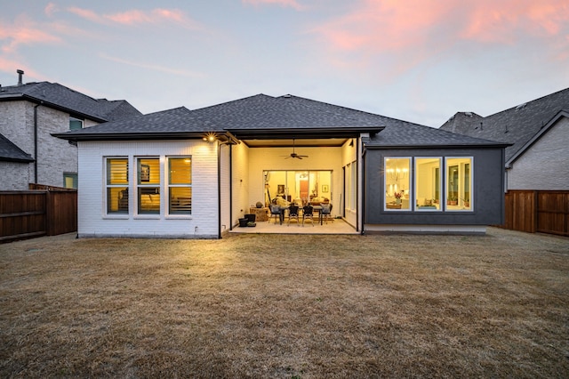 back of house at dusk featuring a shingled roof, a patio, a fenced backyard, ceiling fan, and a yard