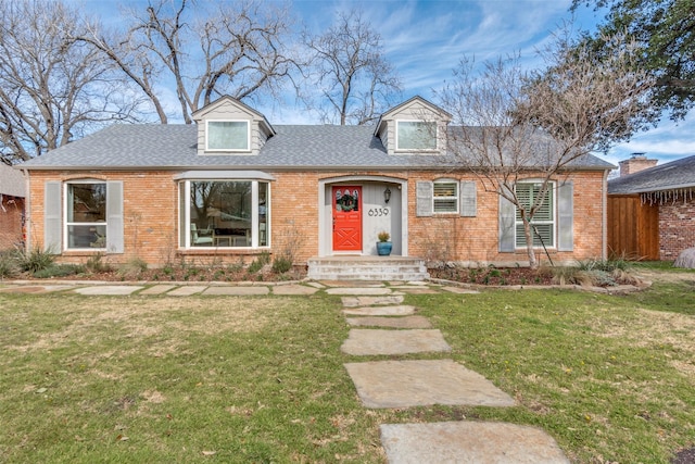 new england style home with a front lawn, roof with shingles, fence, and brick siding